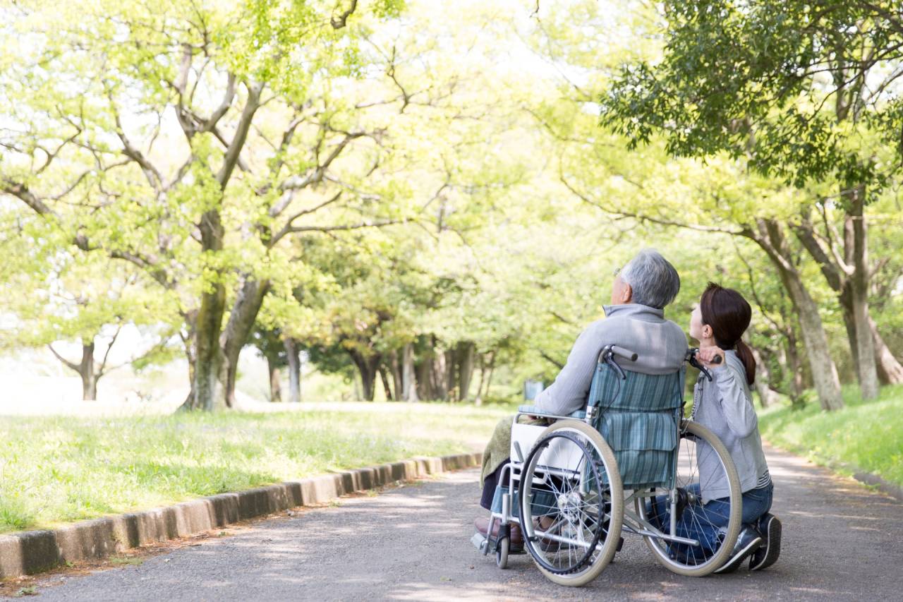 緑豊かな公園で、車いすを利用する高齢の男性と付き添う女性が過ごすイメージ